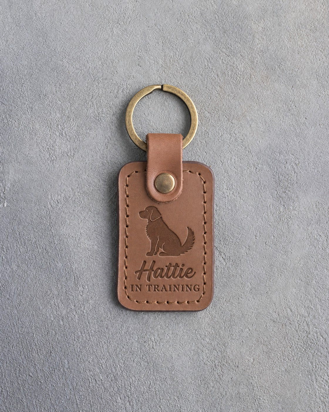 Caramel leather keychain debossed with a small seated puppy above Hattie and IN TRAINING, on gray concrete