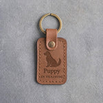 Caramel leather keychain debossed with a small seated puppy above Puppy and IN TRAINING, on gray concrete