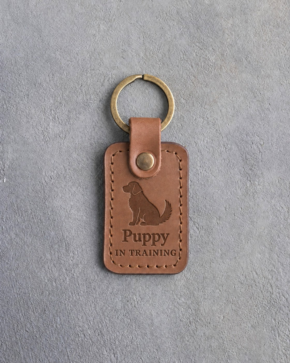 Caramel leather keychain debossed with a small seated puppy above Puppy and IN TRAINING, on gray concrete