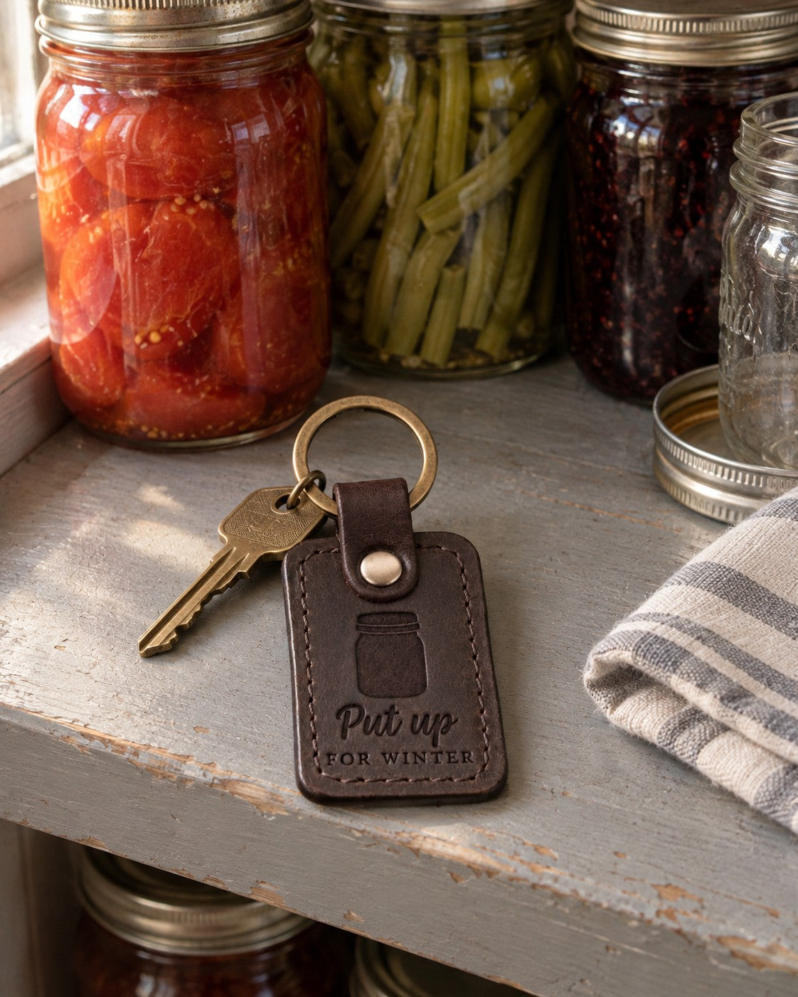 Dark brown Put Up for Winter keychain on a gray painted pantry shelf in front of jars of home-canned tomatoes and beans