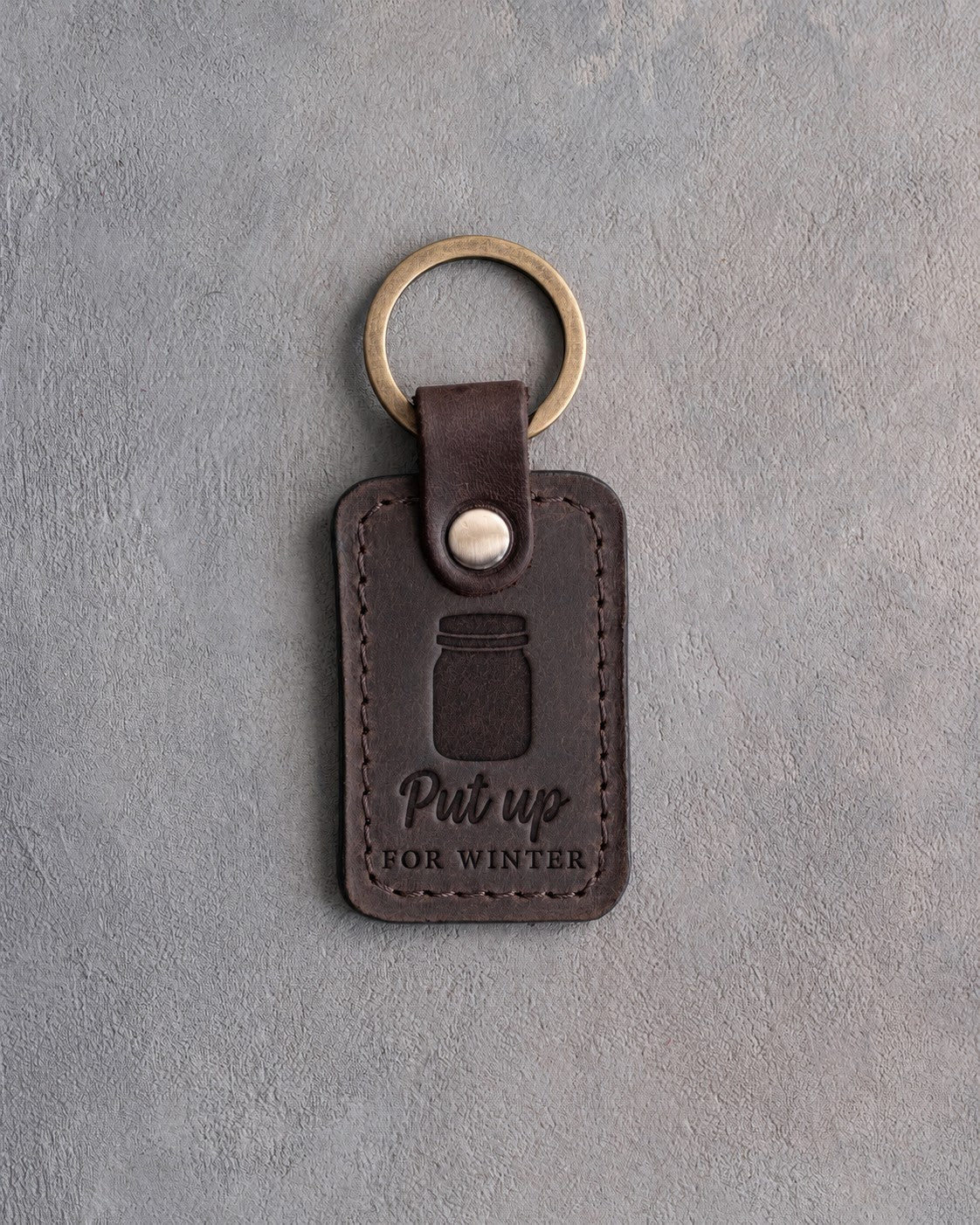 Dark espresso leather keychain debossed with a mason jar above the words Put up FOR WINTER, on gray concrete