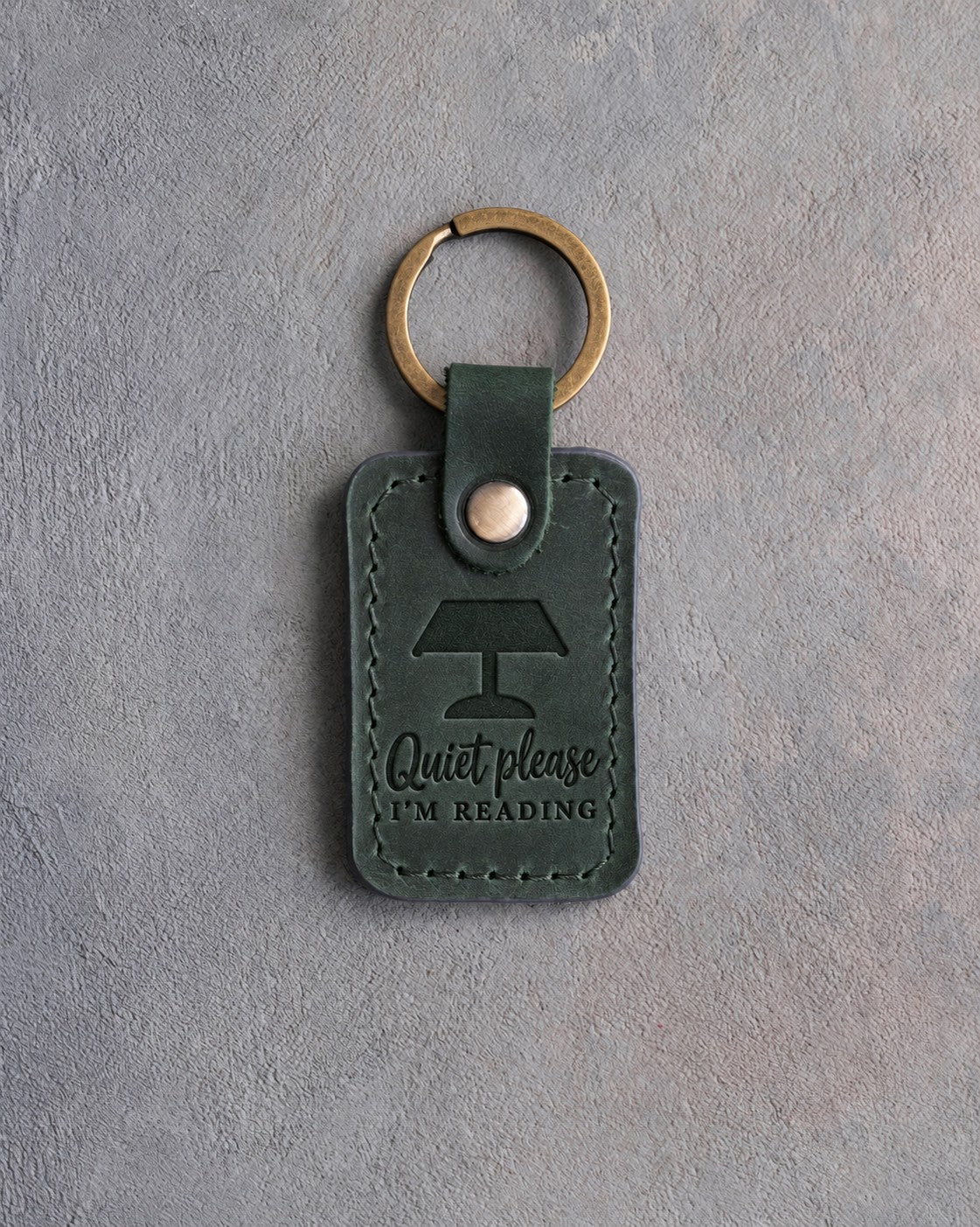 Forest green leather keychain debossed with a banker's lamp above Quiet please and I'M READING, on gray concrete
