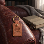 Caramel Raised a READER keychain on a leather armchair arm above a closed book and folded reading glasses