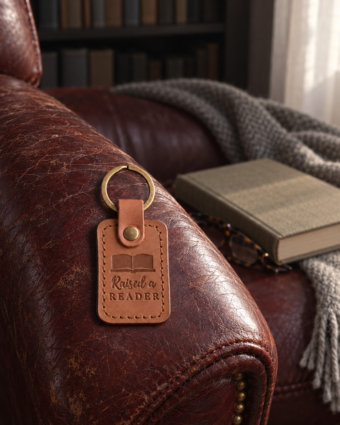 Caramel Raised a READER keychain on a leather armchair arm above a closed book and folded reading glasses