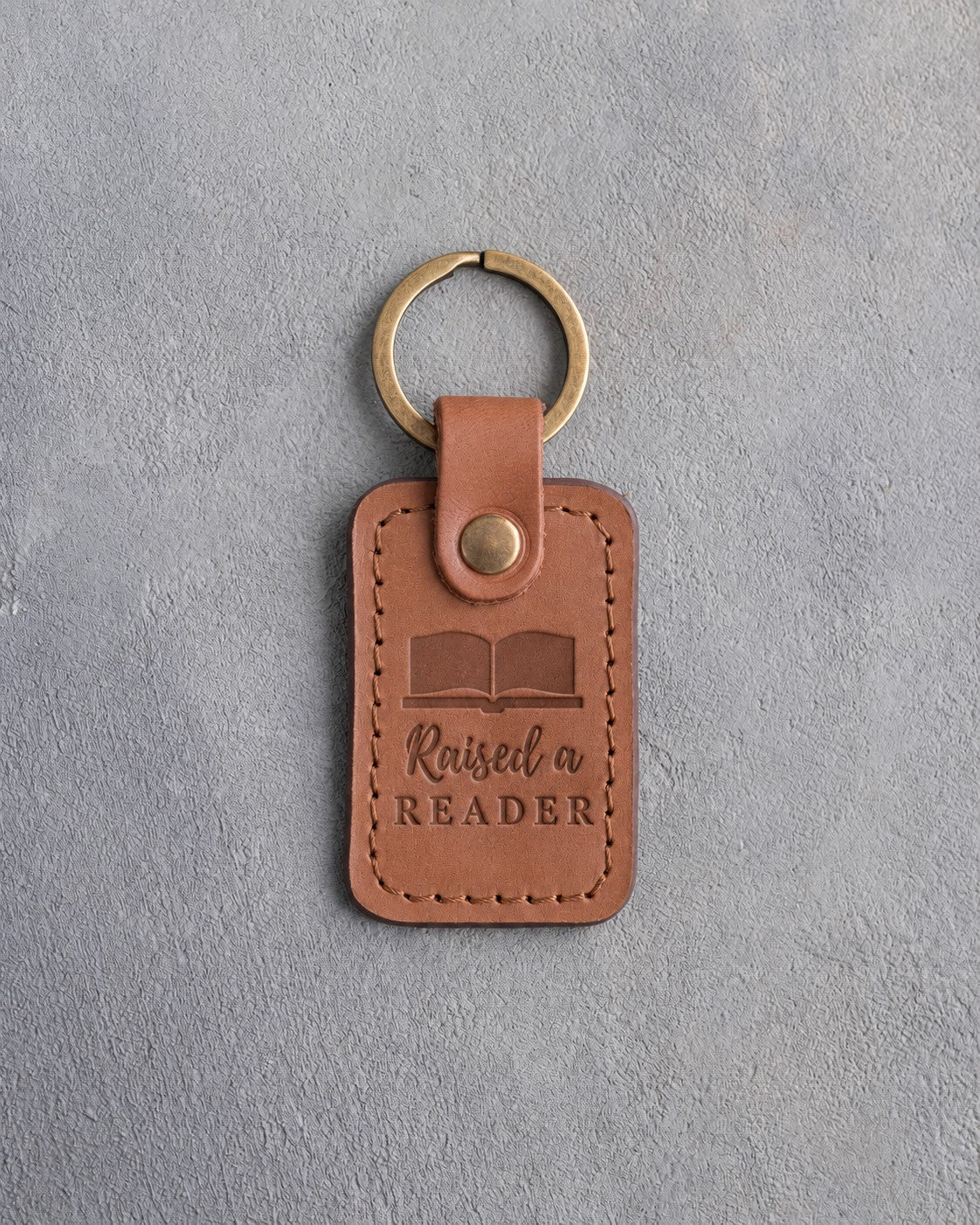 Caramel leather keychain debossed with an open book above Raised a and READER, on gray concrete
