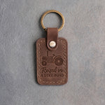 Sicilian brown leather keychain debossed with an open-station tractor above the words Raised on A DIRT ROAD, on gray concrete