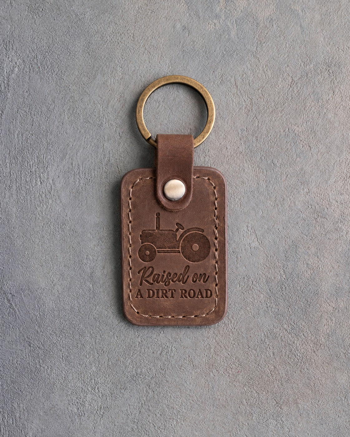 Sicilian brown leather keychain debossed with an open-station tractor above the words Raised on A DIRT ROAD, on gray concrete