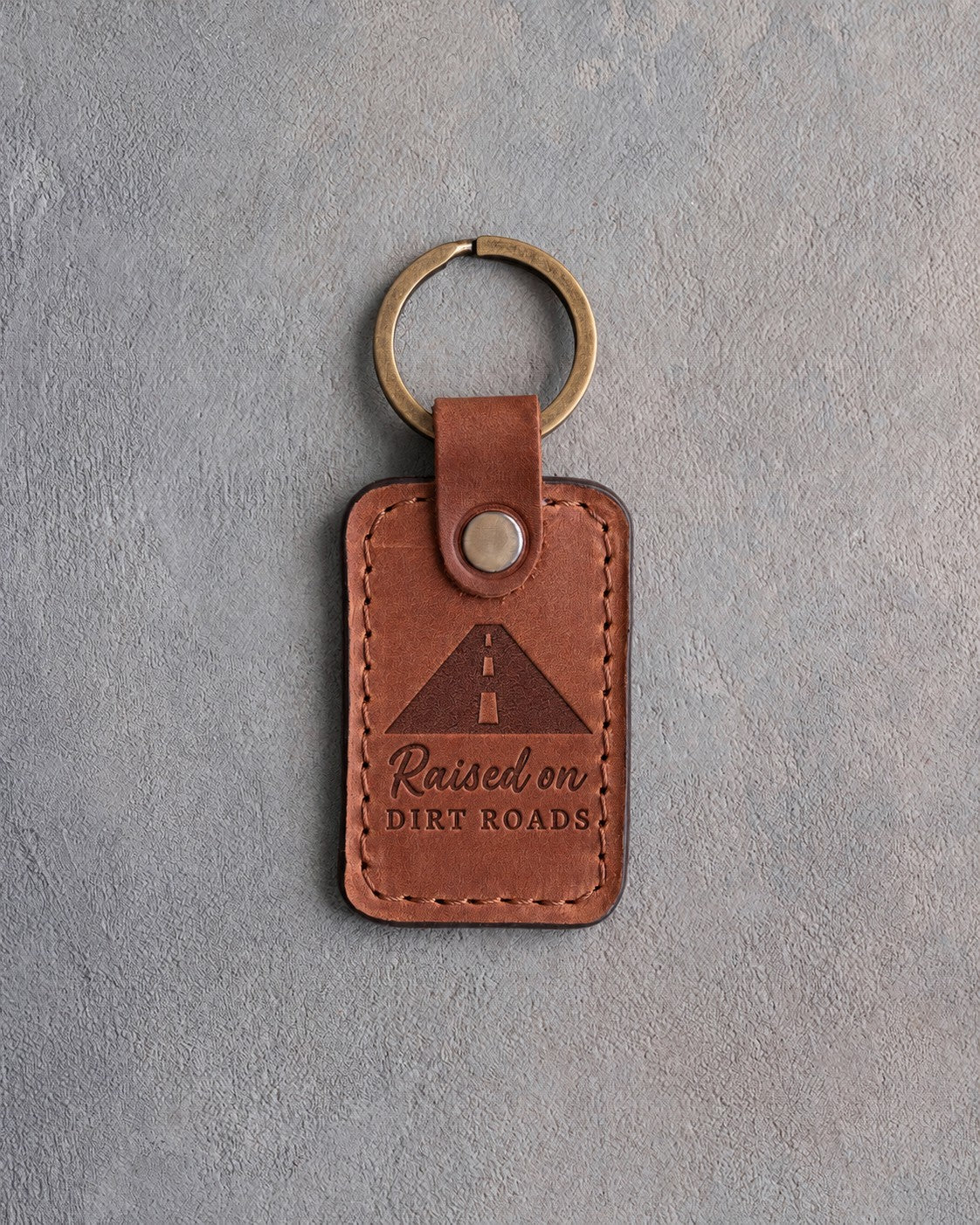 Arizona tan leather keychain debossed with a road to the horizon above Raised on and DIRT ROADS