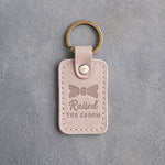 Gray sand leather keychain debossed with a bow tie above the words Raised THE GROOM, on gray concrete