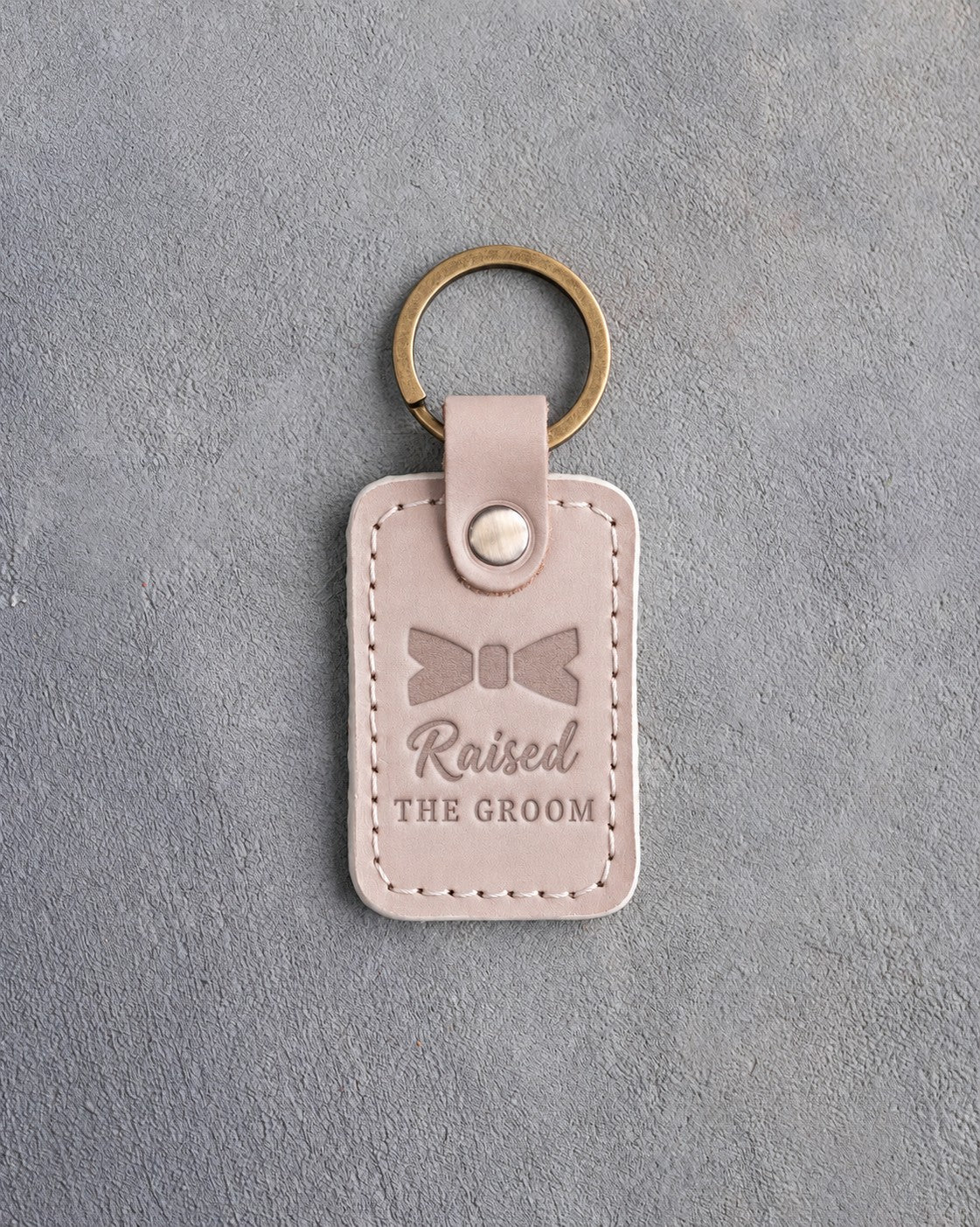 Gray sand leather keychain debossed with a bow tie above the words Raised THE GROOM, on gray concrete