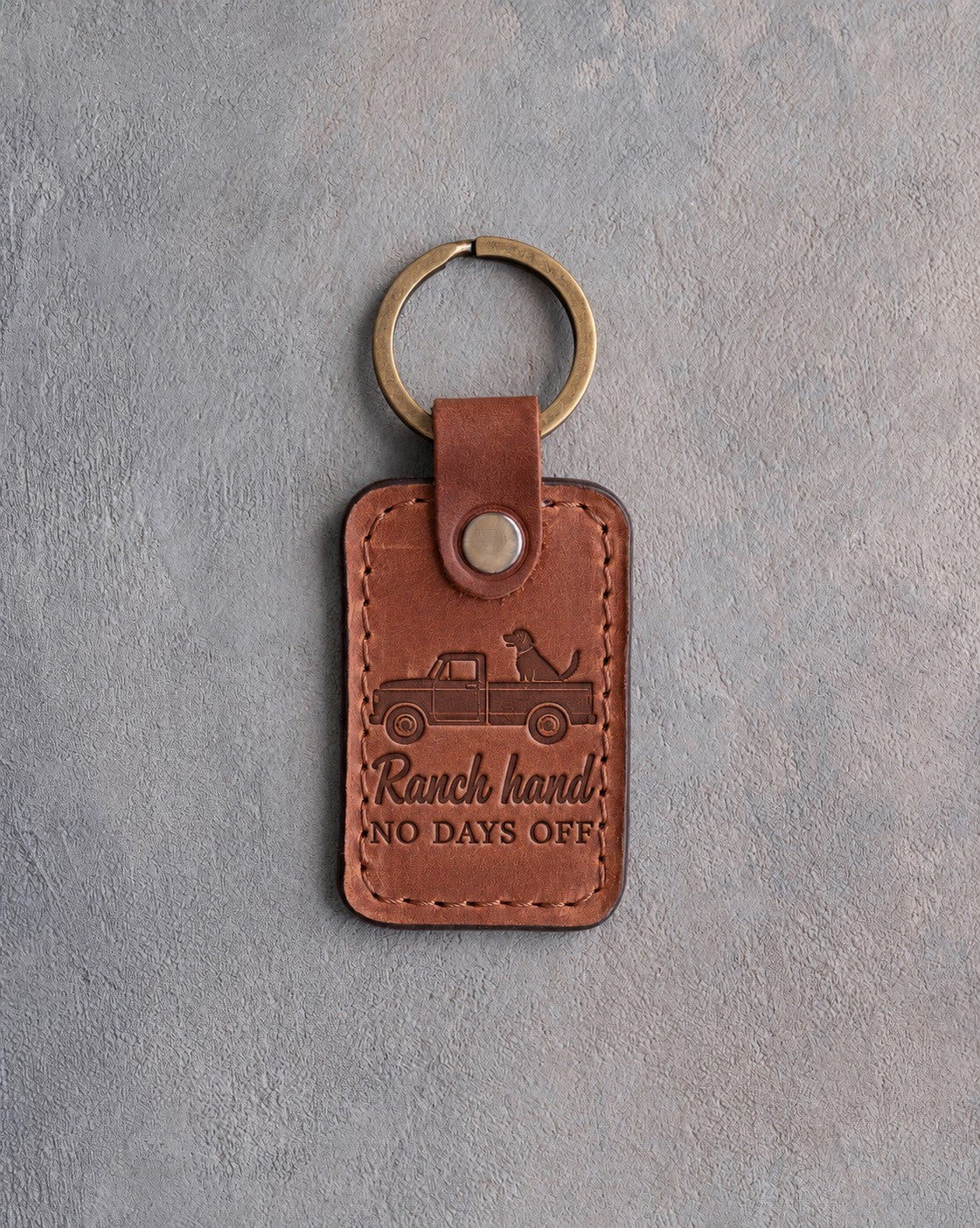Arizona tan leather keychain debossed with a dog riding in a pickup truck bed and the words Ranch hand NO DAYS OFF, on gray concrete