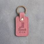 Parisian Blossom pink leather keychain debossed with a cowboy boot above Ranch and MOM, on gray plaster