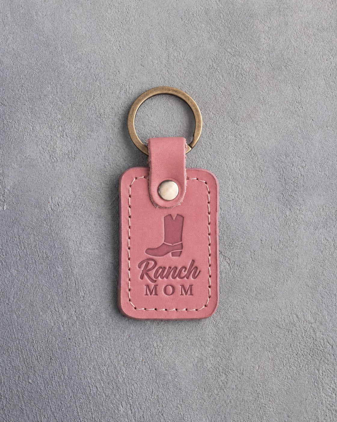 Parisian Blossom pink leather keychain debossed with a cowboy boot above Ranch and MOM, on gray plaster
