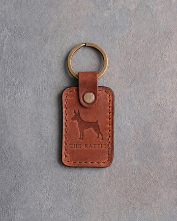 Arizona tan leather keychain debossed with a Rat Terrier silhouette above THE RATTIE, on gray concrete