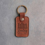 Arizona tan leather keychain debossed with a row of books on a shelf above Reading and SINCE 1987, on gray concrete