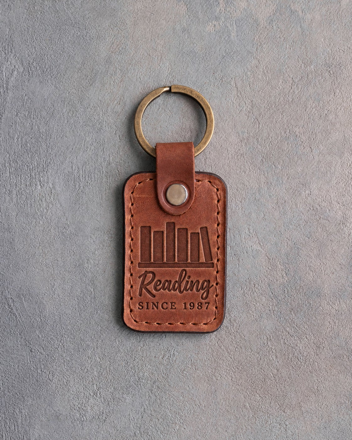 Arizona tan leather keychain debossed with a row of books on a shelf above Reading and SINCE 1987, on gray concrete