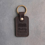 Dark espresso leather keychain debossed with an open book above Reads by and THE FIRE, on gray concrete