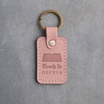 Blush pink leather keychain debossed with an open book above Reads to and OLIVIA, on gray concrete