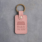 Blush pink leather keychain debossed with an open book above Reads to and THE KIDS, on gray concrete