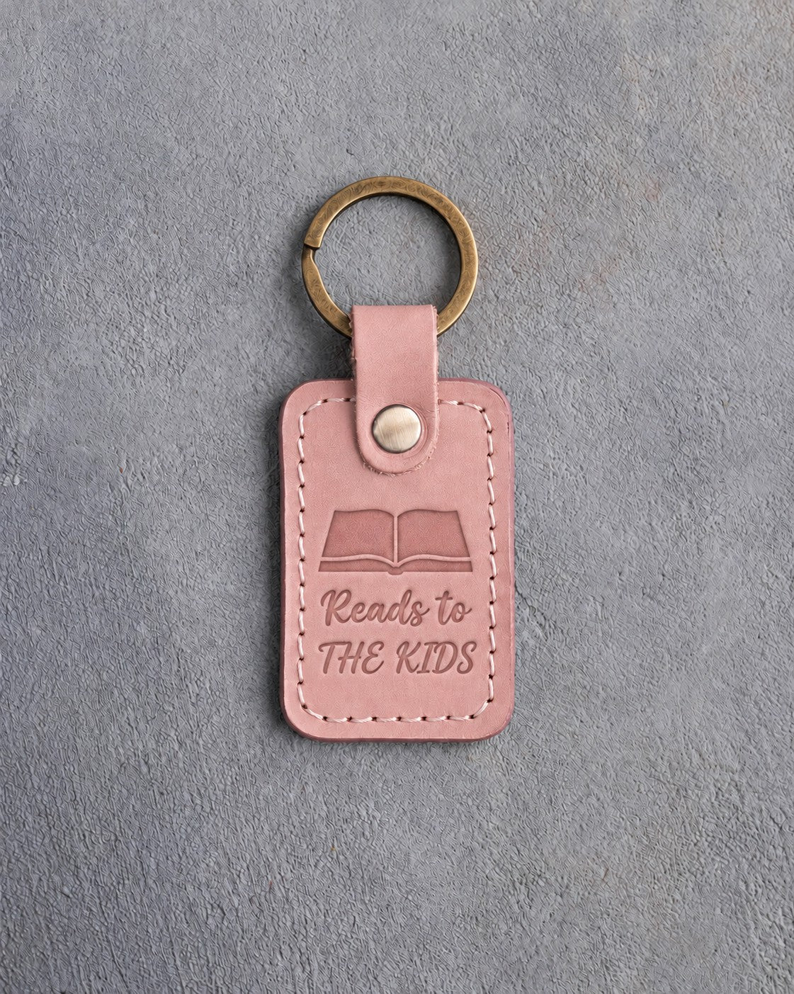 Blush pink leather keychain debossed with an open book above Reads to and THE KIDS, on gray concrete