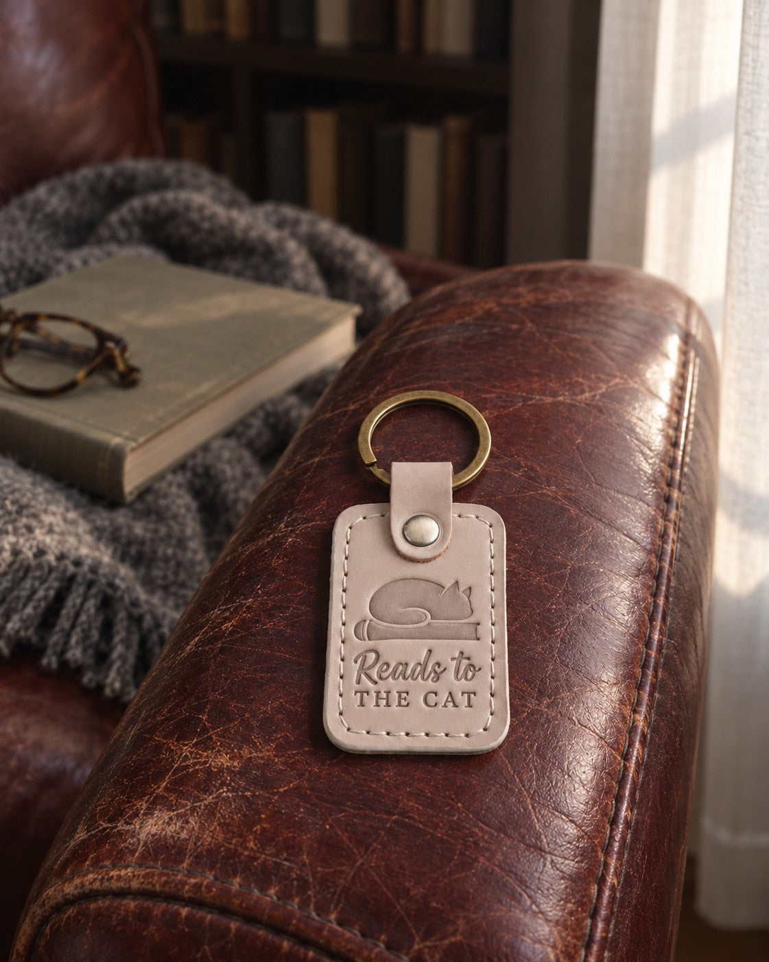Pale gray Reads to THE CAT keychain on an armchair arm with a tabby cat asleep on the cushion behind