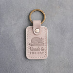 Gray sand leather keychain debossed with a cat asleep on a book above Reads to and THE CAT, on gray concrete