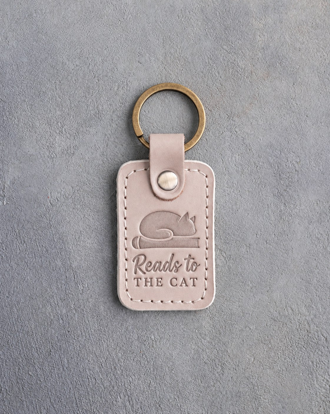 Gray sand leather keychain debossed with a cat asleep on a book above Reads to and THE CAT, on gray concrete