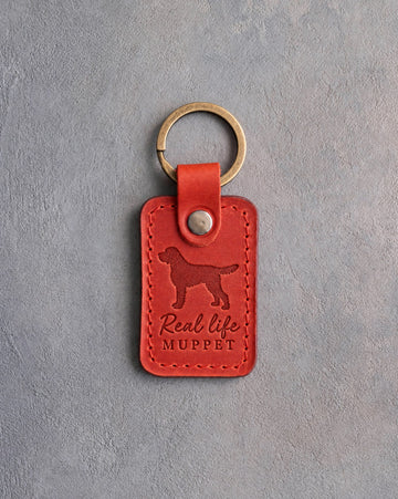 Poppy red leather keychain debossed with a Labradoodle silhouette and the words Real life MUPPET, on gray concrete
