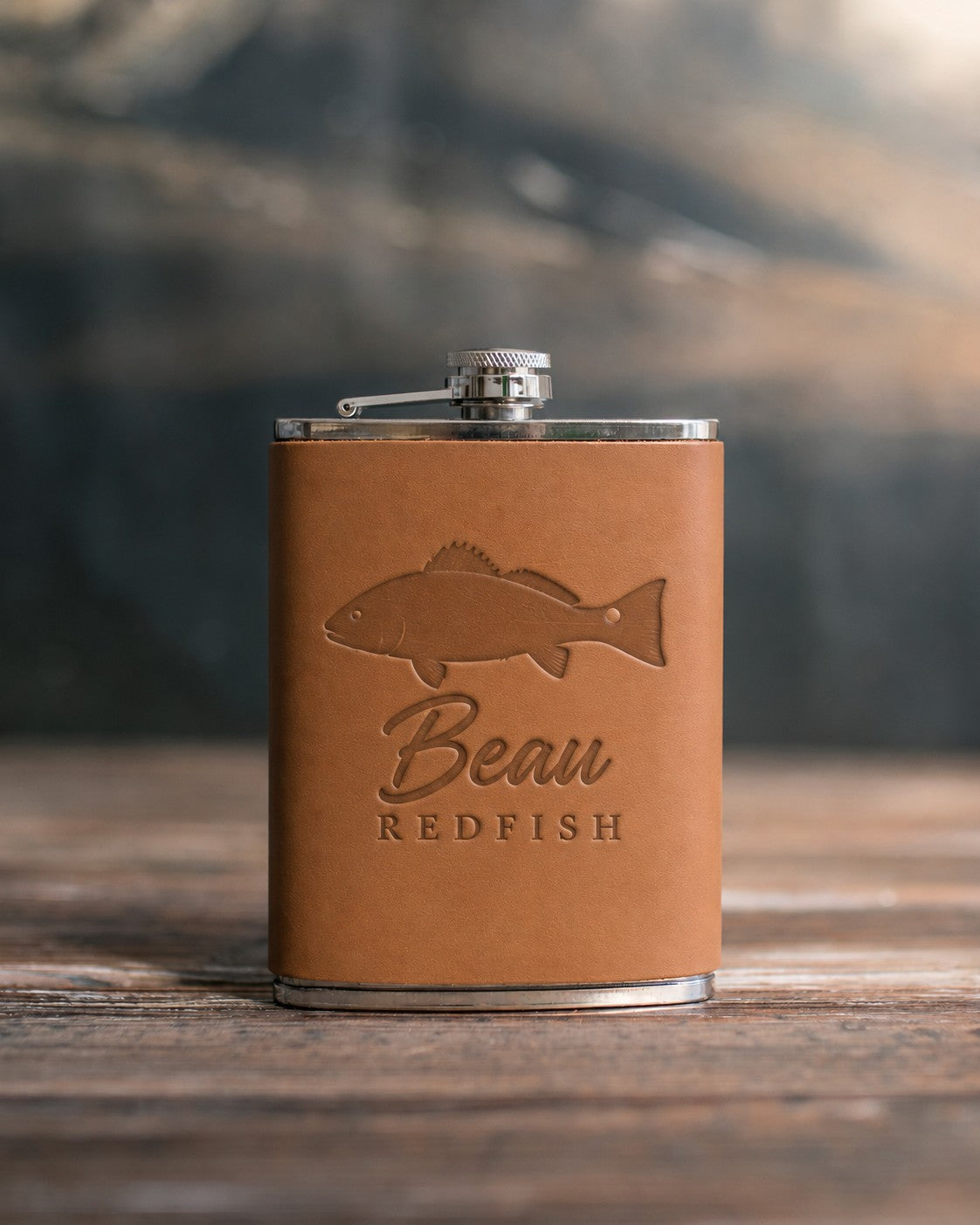 Caramel leather-wrapped hip flask debossed with a redfish above Beau and REDFISH, on a wooden table