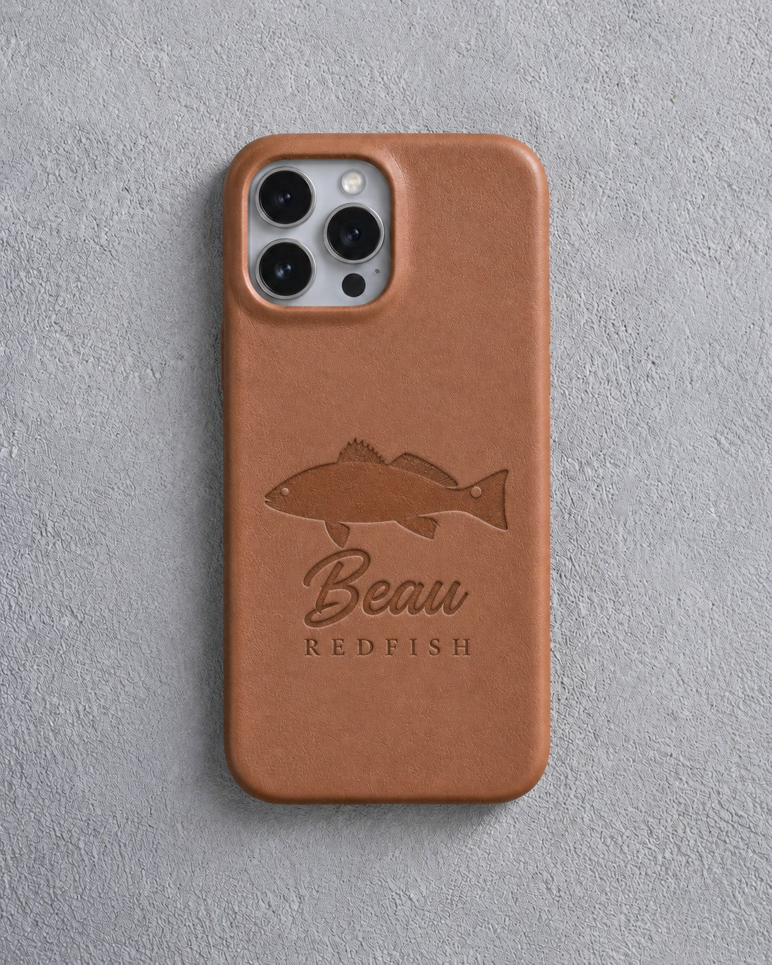 Back of a caramel leather iPhone case debossed with a redfish above Beau and REDFISH below the camera, on gray plaster