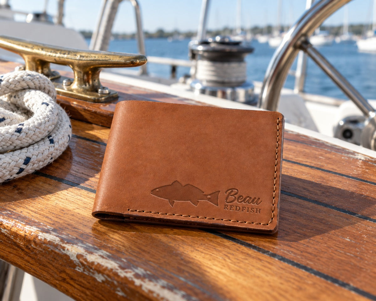 Caramel Beau REDFISH bifold wallet in full-grain leather on the worn teak cockpit table of a sailboat beside a coiled white dock line