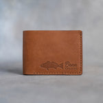 Closed caramel leather bifold wallet with a redfish beside Beau and REDFISH pressed in the lower right corner, on gray plaster