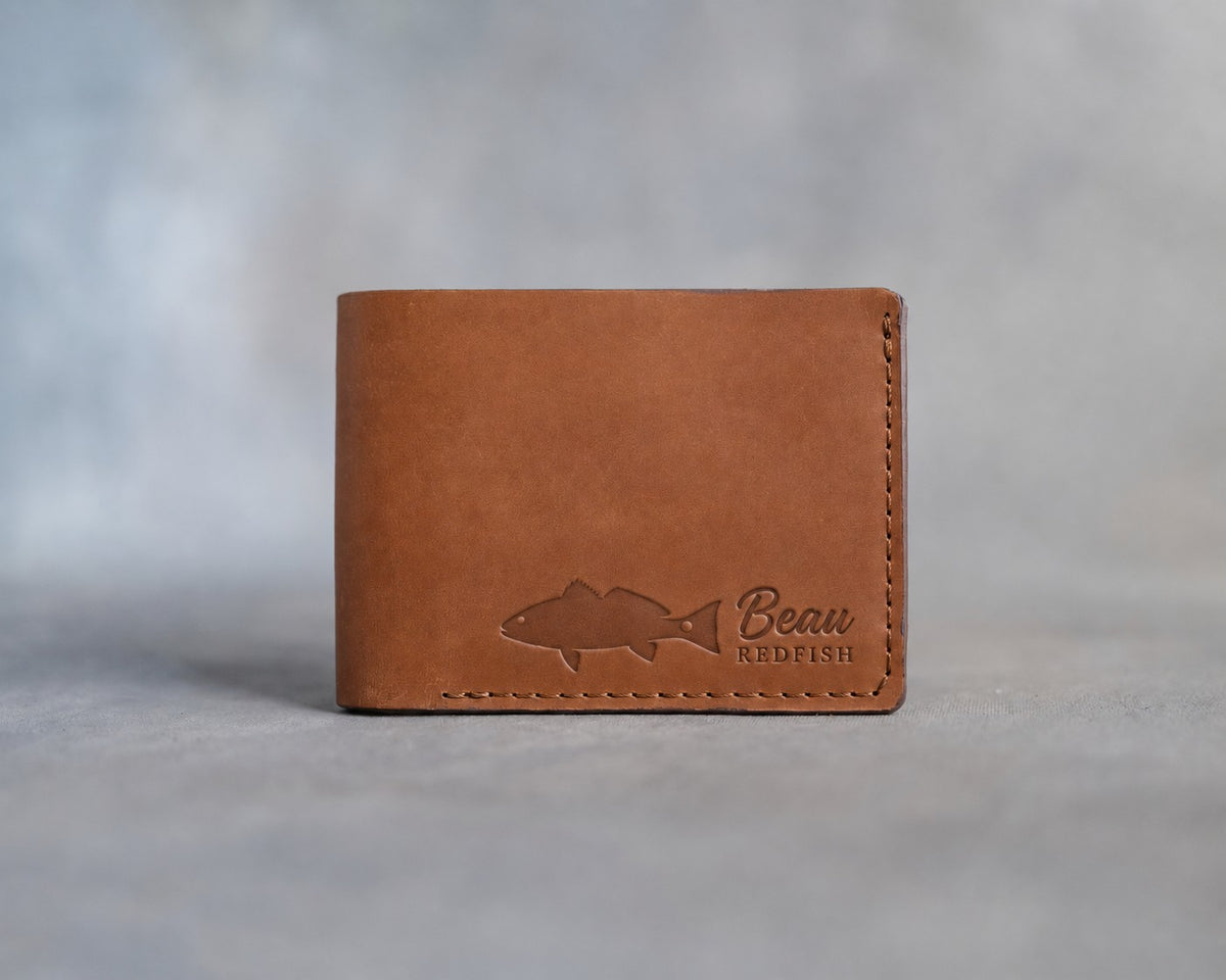 Closed caramel leather bifold wallet with a redfish beside Beau and REDFISH pressed in the lower right corner, on gray plaster