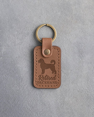 Caramel leather keychain debossed with a Portuguese water dog and the words Retired DECKHAND, on gray concrete