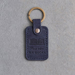 Blueberry navy leather keychain debossed with an open book above Please return and MY BOOKS, on gray concrete
