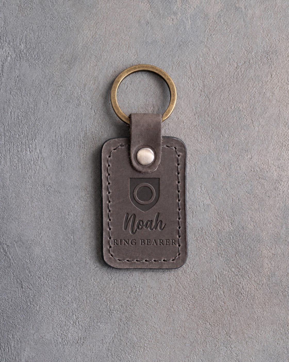 Country gray leather keychain debossed with a shield badge holding a wedding band, above the name Noah and RING BEARER, on gray concrete