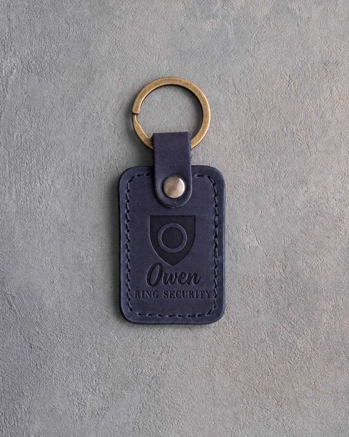Blueberry navy leather keychain debossed with a shield badge holding a wedding band, above the name Owen and RING SECURITY, on gray concrete