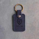 Blueberry navy leather keychain debossed with a shield badge holding a wedding band, above Official and RING SECURITY, on gray concrete