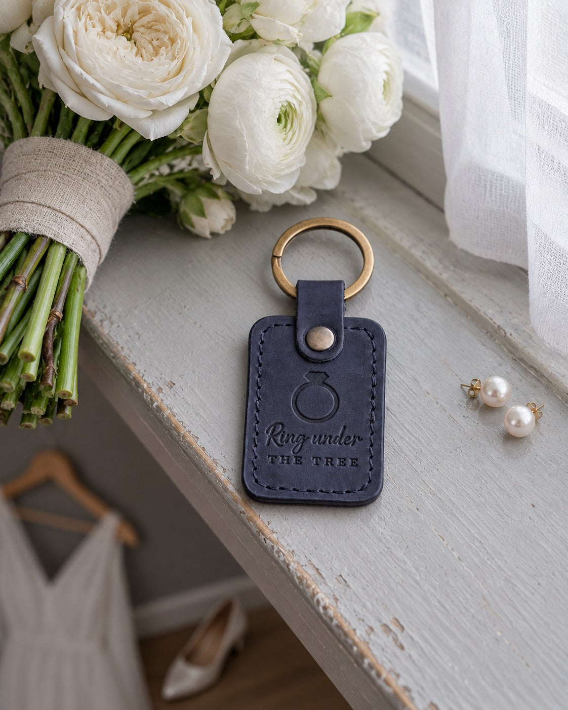 Ring under THE TREE keychain in navy leather on a gray windowsill beside pearl earrings and a hand-tied bouquet