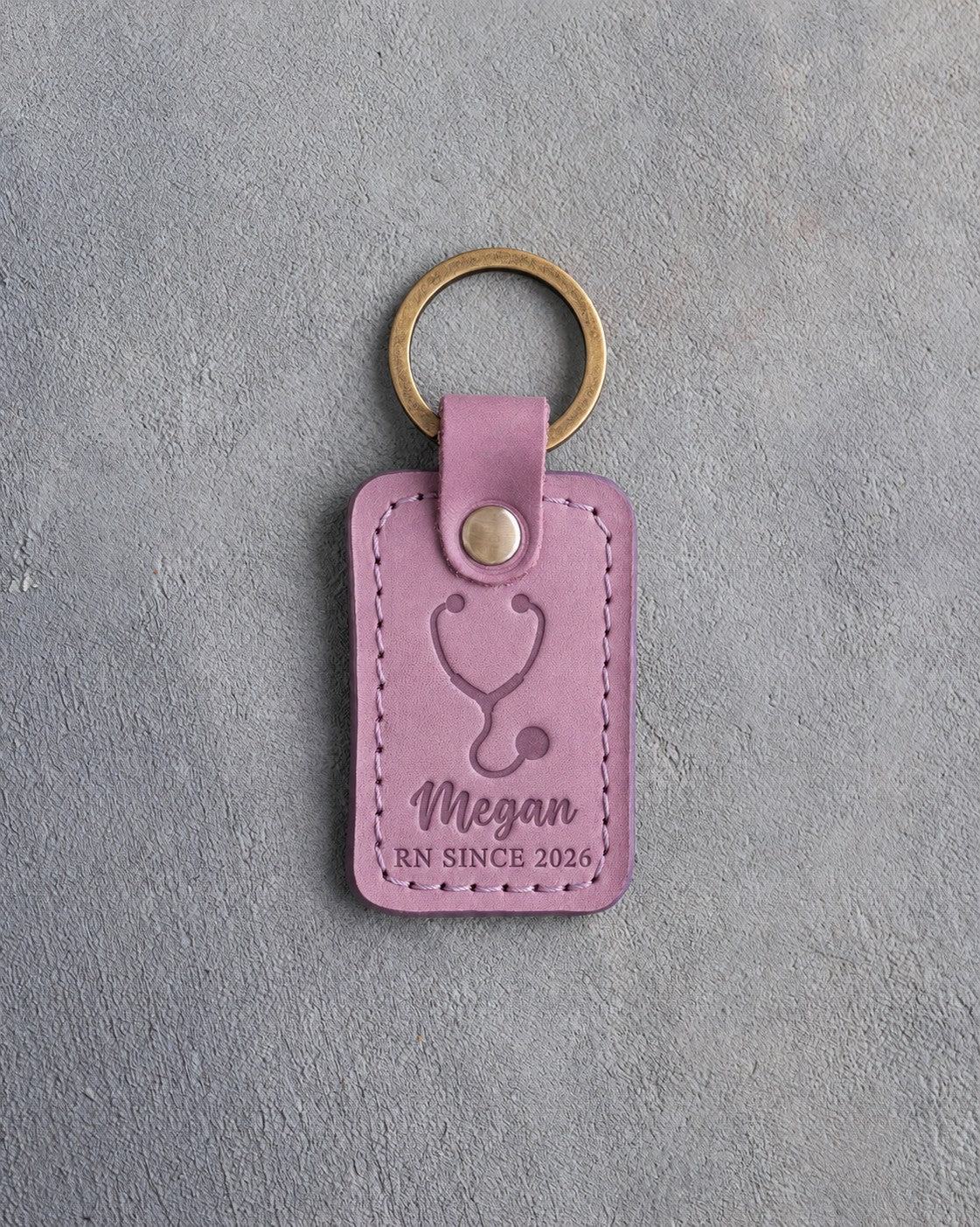 Lilac leather keychain debossed with a stethoscope above the name Megan and RN SINCE 2026, on gray plaster