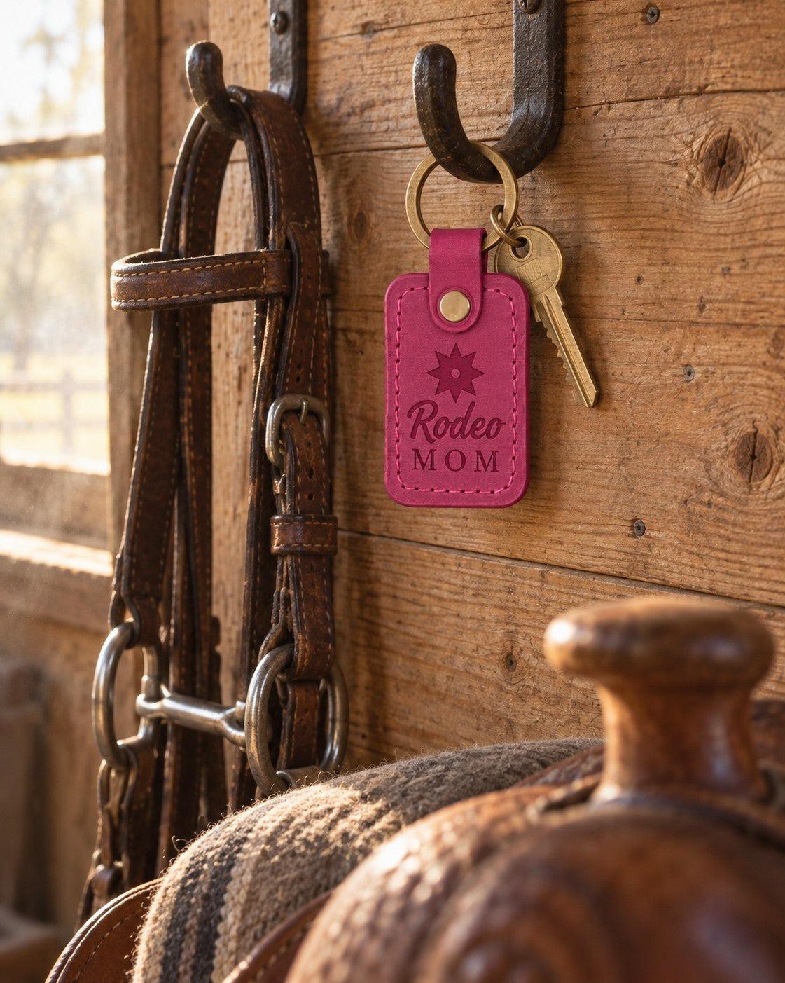 Fuchsia Rodeo MOM keychain hanging from an iron bridle hook on a tack room wall beside a leather headstall