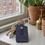 Navy Room for One More keychain on a crowded windowsill among small terracotta pots of houseplants