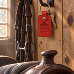 Poppy red Runs the BARN keychain hanging from a forged bridle hook on a pine tack room wall beside a worn headstall