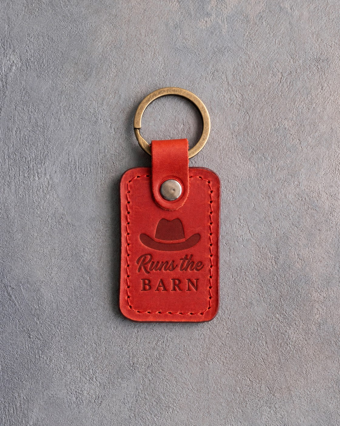 Poppy red leather keychain debossed with a cowboy hat above Runs the and BARN, on gray plaster