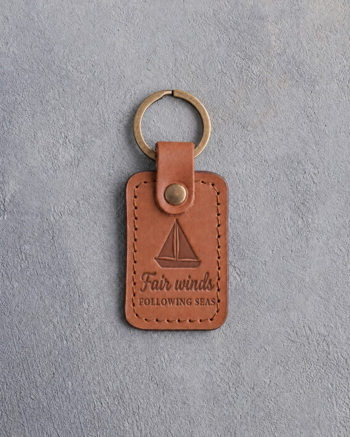Caramel leather keychain debossed with a sloop under full sail above Fair winds and FOLLOWING SEAS, on gray concrete