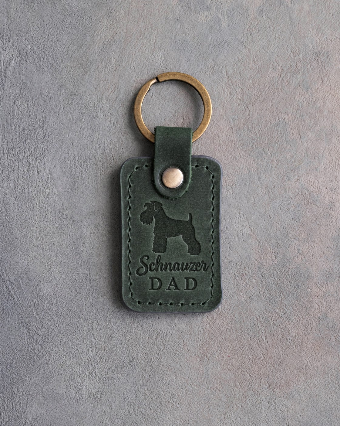 Forest green leather keychain debossed with a Miniature Schnauzer silhouette and the words Schnauzer DAD, on gray concrete