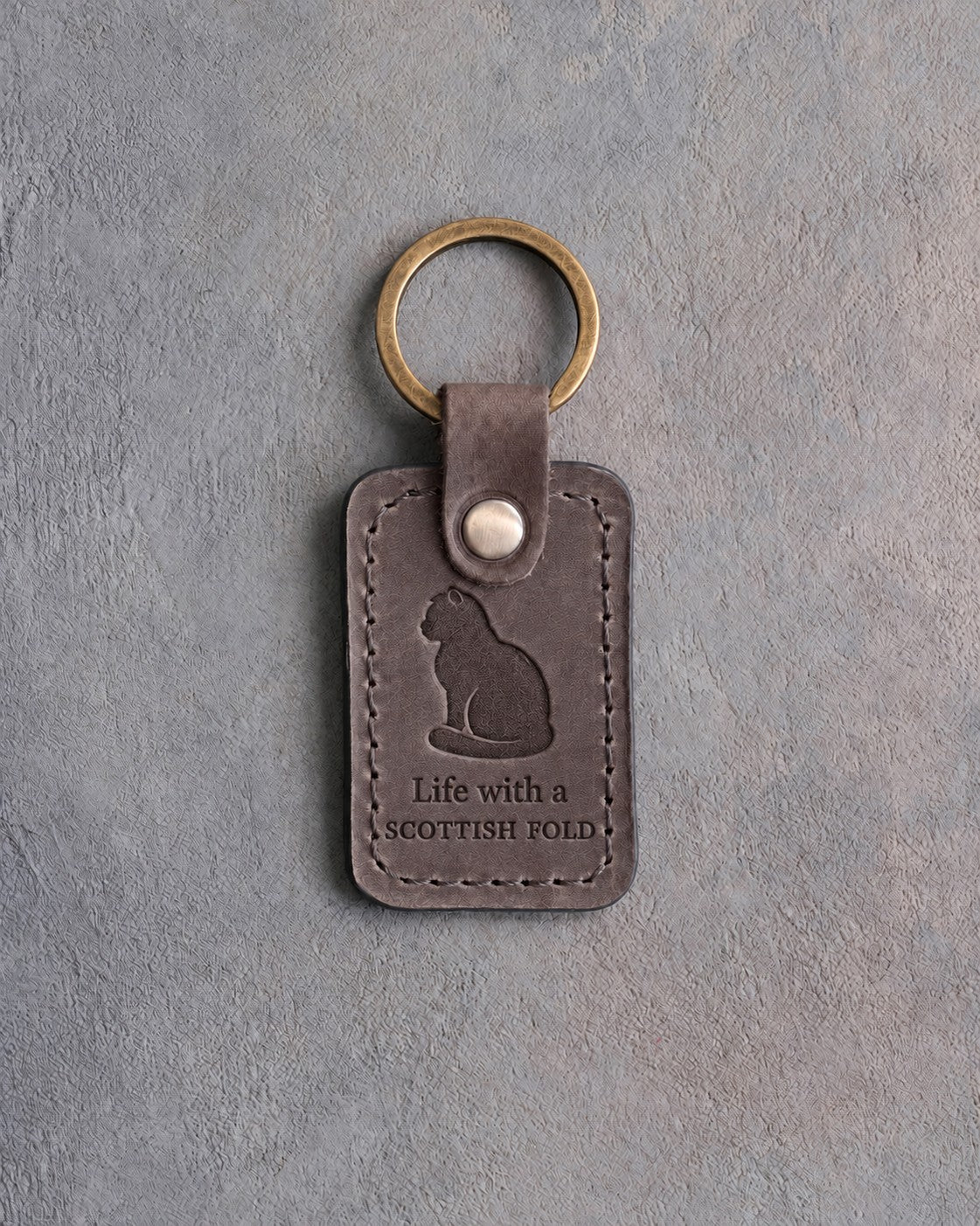 Country gray leather keychain debossed with a Scottish Fold sitting upright above Life with a and SCOTTISH FOLD, on gray concrete