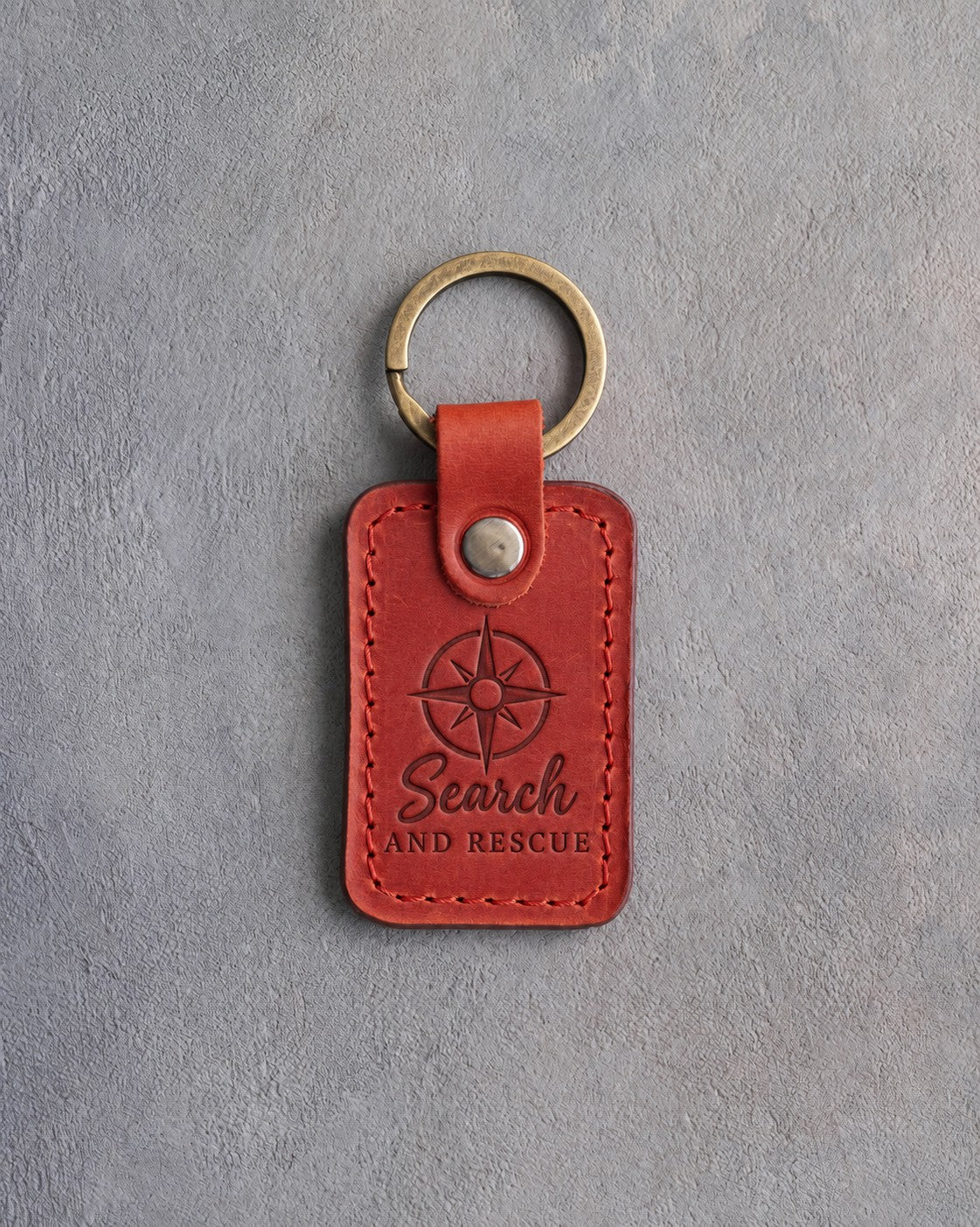 Poppy red leather keychain debossed with a compass rose and the words Search AND RESCUE, on gray concrete