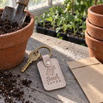 Gray sand Seed Saver keychain on a wooden table beside labeled paper envelopes of dried bean seeds