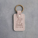 Gray sand leather keychain debossed with an open seed packet above Seed SAVER, on gray concrete